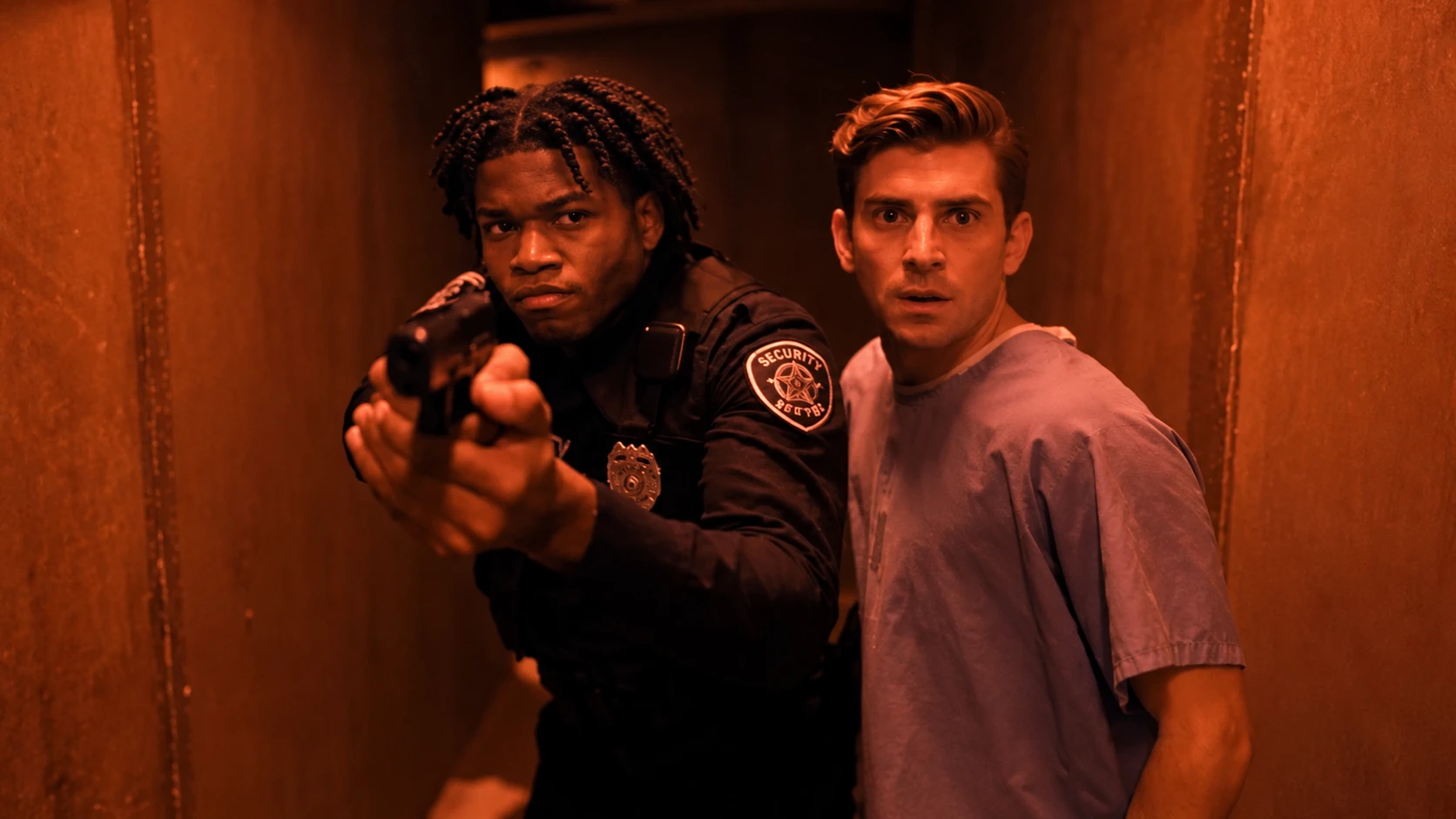 A security officer aims a gun down a red-lit corridor while a man in a hospital gown looks on, startled